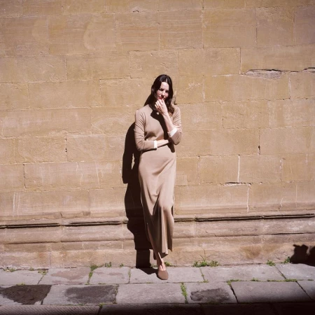 A woman in a tan dress stands against a textured wall. She is in full shot with her hand near her face.