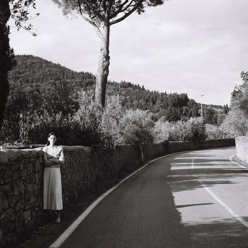 Monochrome portrait of a woman in a dress standing against a stone wall along a rural road, with trees and hills in background.