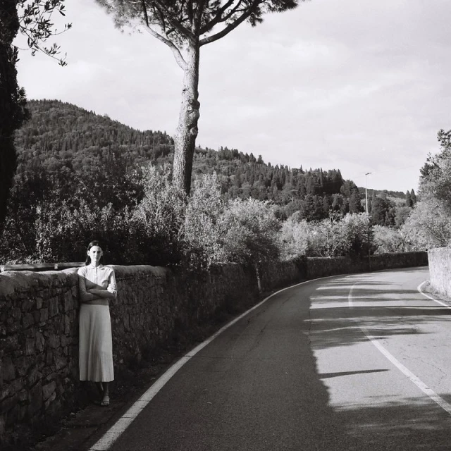 Monochrome portrait of a woman in a dress standing against a stone wall along a rural road, with trees and hills in background.