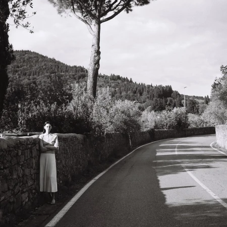 Monochrome portrait of a woman in a dress standing against a stone wall along a rural road, with trees and hills in background.