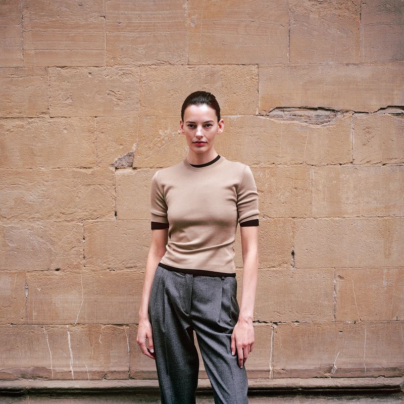 A slender woman poses in a short-sleeved top and pleated pants, standing against a stone wall backdrop in soft, natural light.