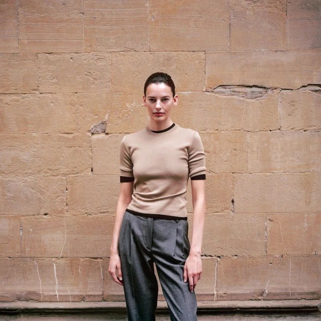 A slender woman poses in a short-sleeved top and pleated pants, standing against a stone wall backdrop in soft, natural light.
