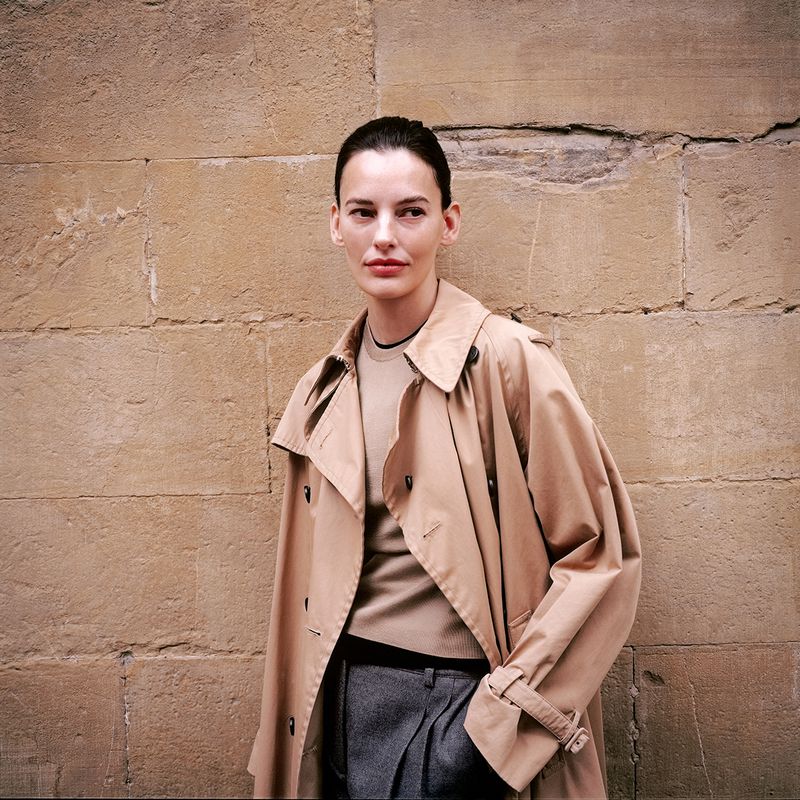 Woman in beige trench coat stands against a stone wall in medium shot; neutral fashion, natural light, and modern styling are visible.