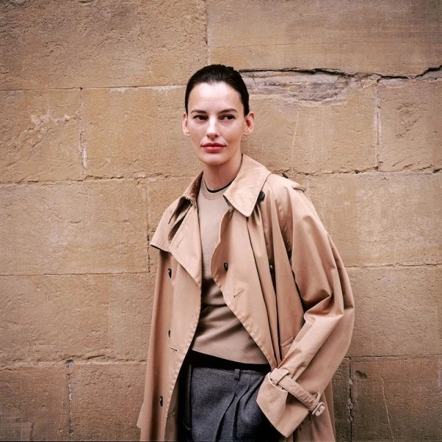 Woman in beige trench coat stands against a stone wall in medium shot; neutral fashion, natural light, and modern styling are visible.