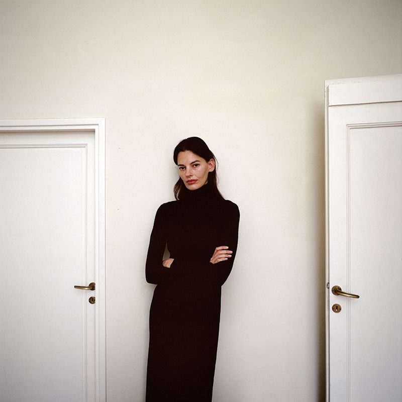 Woman in black turtleneck dress posing between two white doors. Portrait photography with simple, contrasting color palette. Fashion editorial.