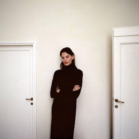 Woman in black turtleneck dress posing between two white doors. Portrait photography with simple, contrasting color palette. Fashion editorial.