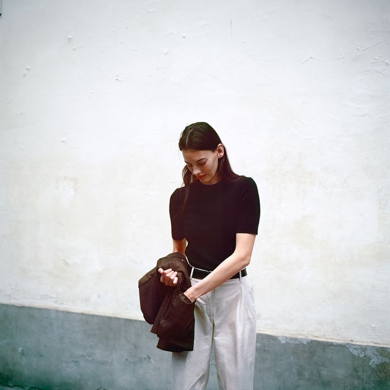A woman in a black top and light pants stands against a textured white wall, holding a brown jacket and placing her hand in her pocket.