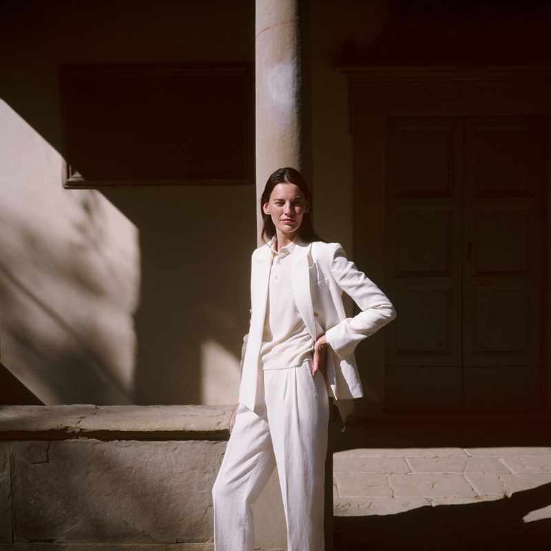 Woman in white suit standing in courtyard with architectural columns, bathed in warm light and shadows. Fashion editorial shoot.