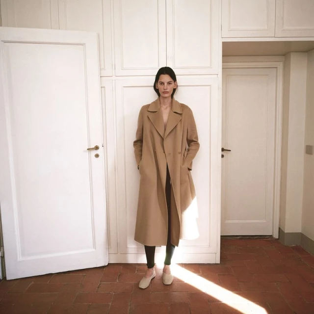 A tall woman with long dark hair stands in a long tan coat with her hands in the pockets, posing in front of white doors indoors.