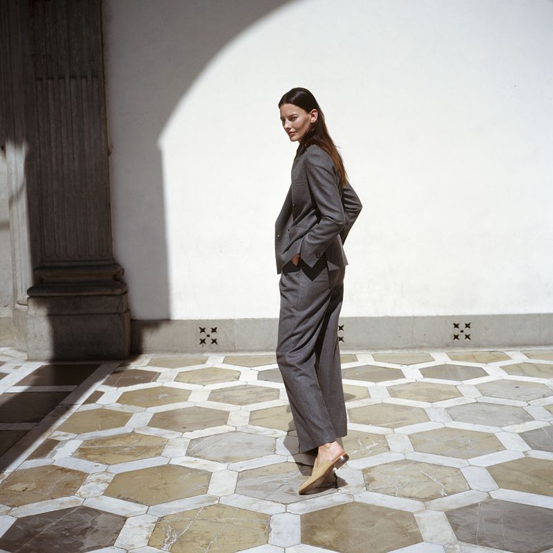 Adult female in grey suit stands in courtyard with geometric tile floor, white walls and arched opening in background.