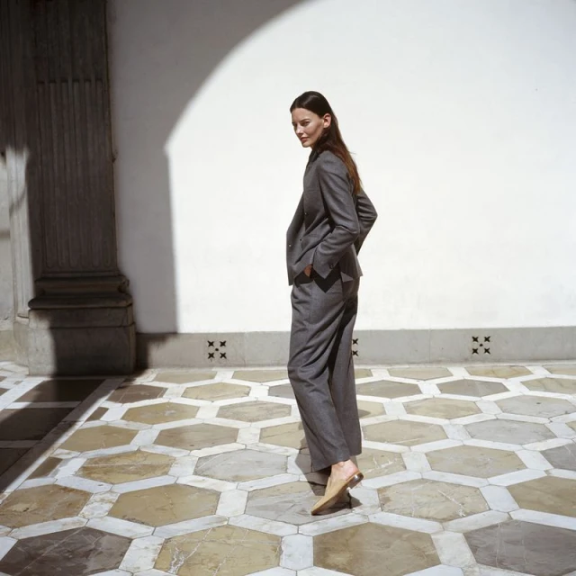 Adult female in grey suit stands in courtyard with geometric tile floor, white walls and arched opening in background.