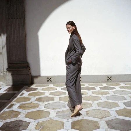 Adult female in grey suit stands in courtyard with geometric tile floor, white walls and arched opening in background.