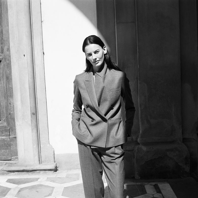 A woman poses in a black and white shot, wearing a gray suit with a double-breasted blazer, standing on a stone-tiled surface.