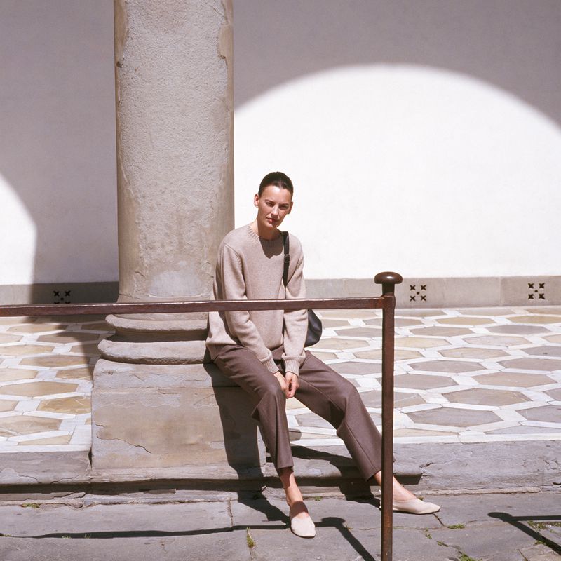 Woman in casual clothes sits on steps next to a stone column in a sunlit courtyard, wearing a sweater, pants, and loafers.
