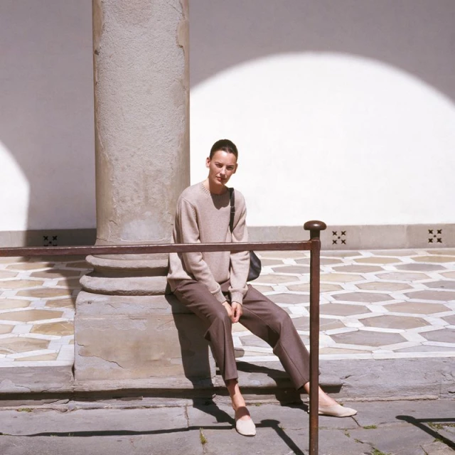 Woman in casual clothes sits on steps next to a stone column in a sunlit courtyard, wearing a sweater, pants, and loafers.