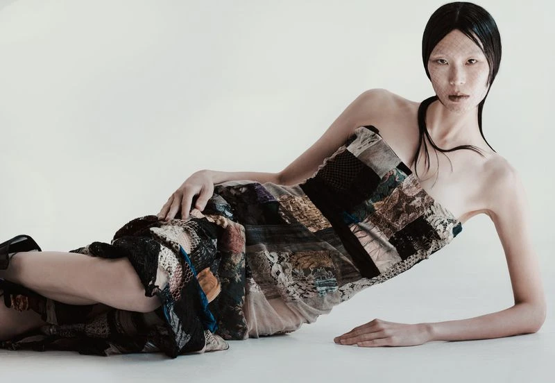 Asian female model lying down in a patchwork dress with white net over her face in a studio setting with even lighting.