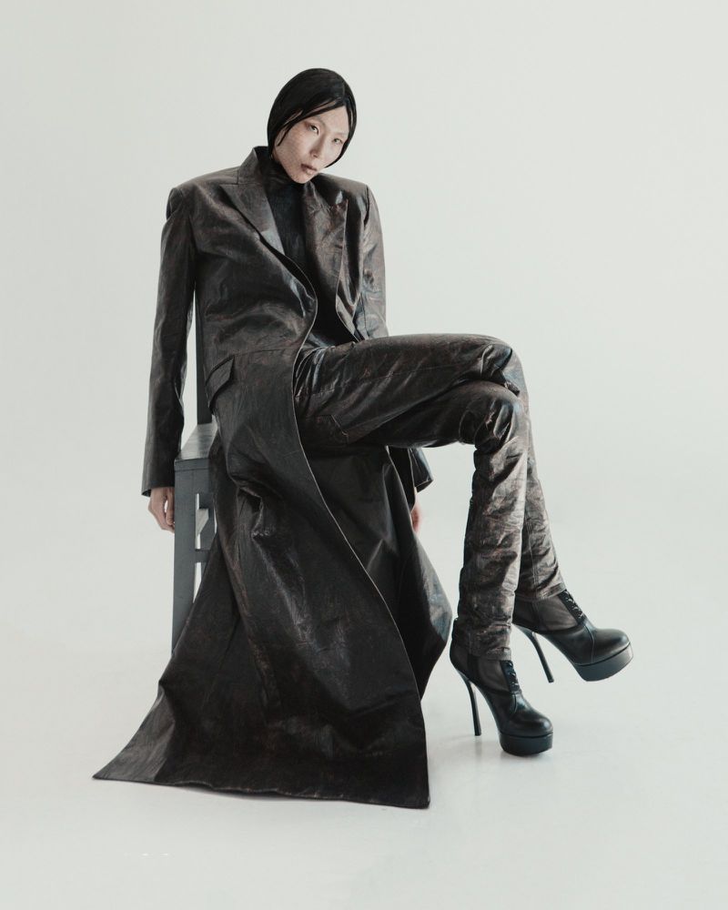 A slender Asian woman with sleek hair poses in a long, textured brown coat, matching pants, and high-heeled boots, seated on a gray stool.
