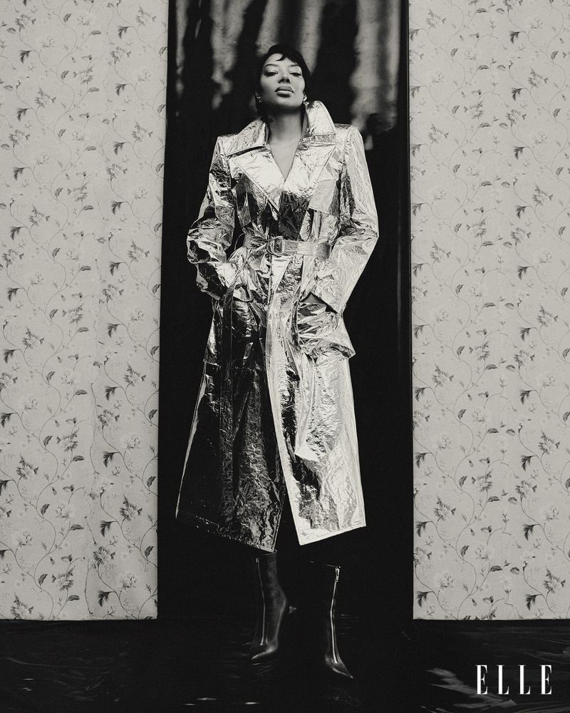 Woman in silver trench coat poses against patterned wallpaper and dark backdrop, creating a high-contrast fashion editorial image.