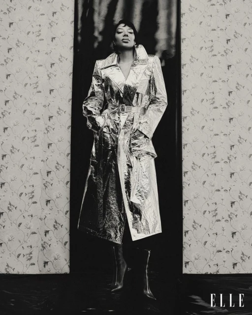 Woman in silver trench coat poses against patterned wallpaper and dark backdrop, creating a high-contrast fashion editorial image.