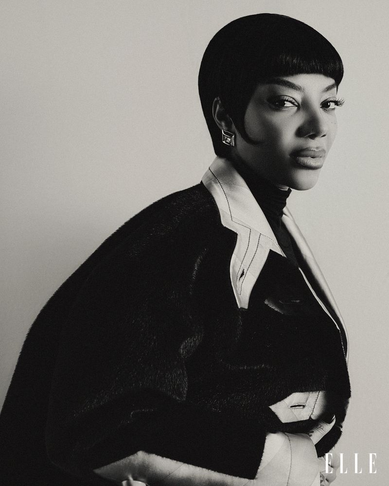 Monochrome portrait of a woman with short black hair wearing a jacket; an ELLE magazine logo is visible in the lower right corner.