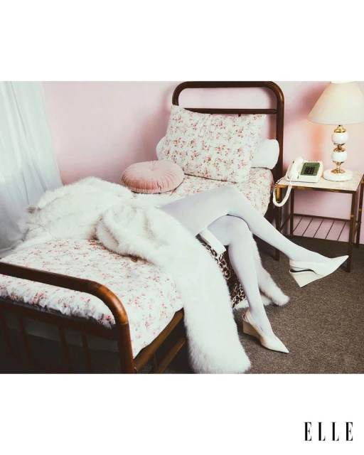 A bed with floral bedding and a white fur coat. A person in white tights and shoes sits on the bed. A telephone sits on a bedside table.