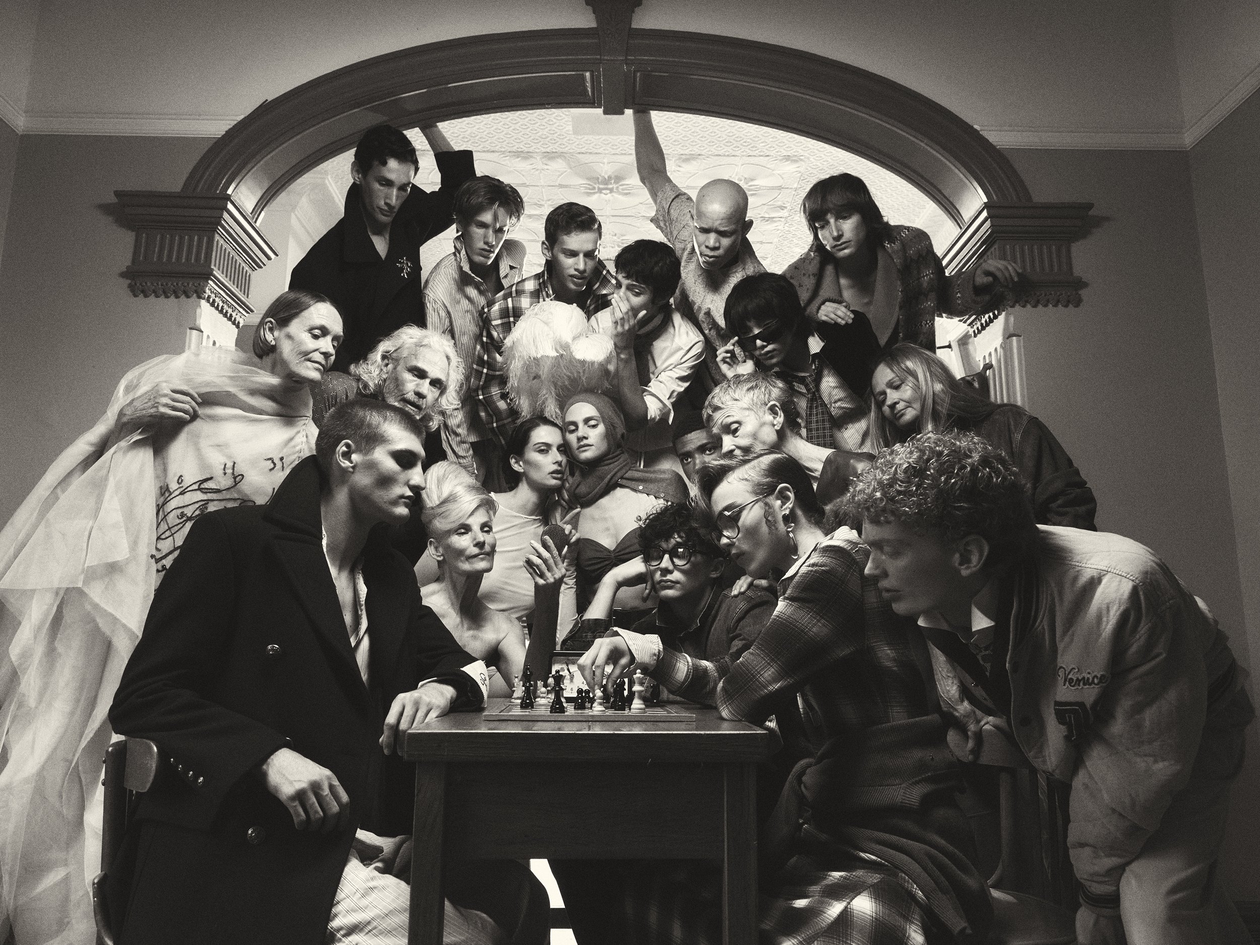 Monochrome studio portrait features a large group of diverse individuals gathered around a chess table in an ornate indoor setting.