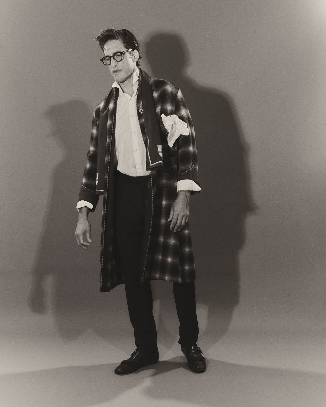 Monochrome portrait of adult male wearing glasses, white button-down shirt, plaid robe, dark pants and leather shoes standing indoors.