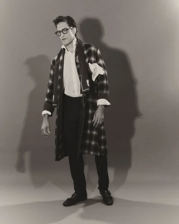 Monochrome portrait of adult male wearing glasses, white button-down shirt, plaid robe, dark pants and leather shoes standing indoors.