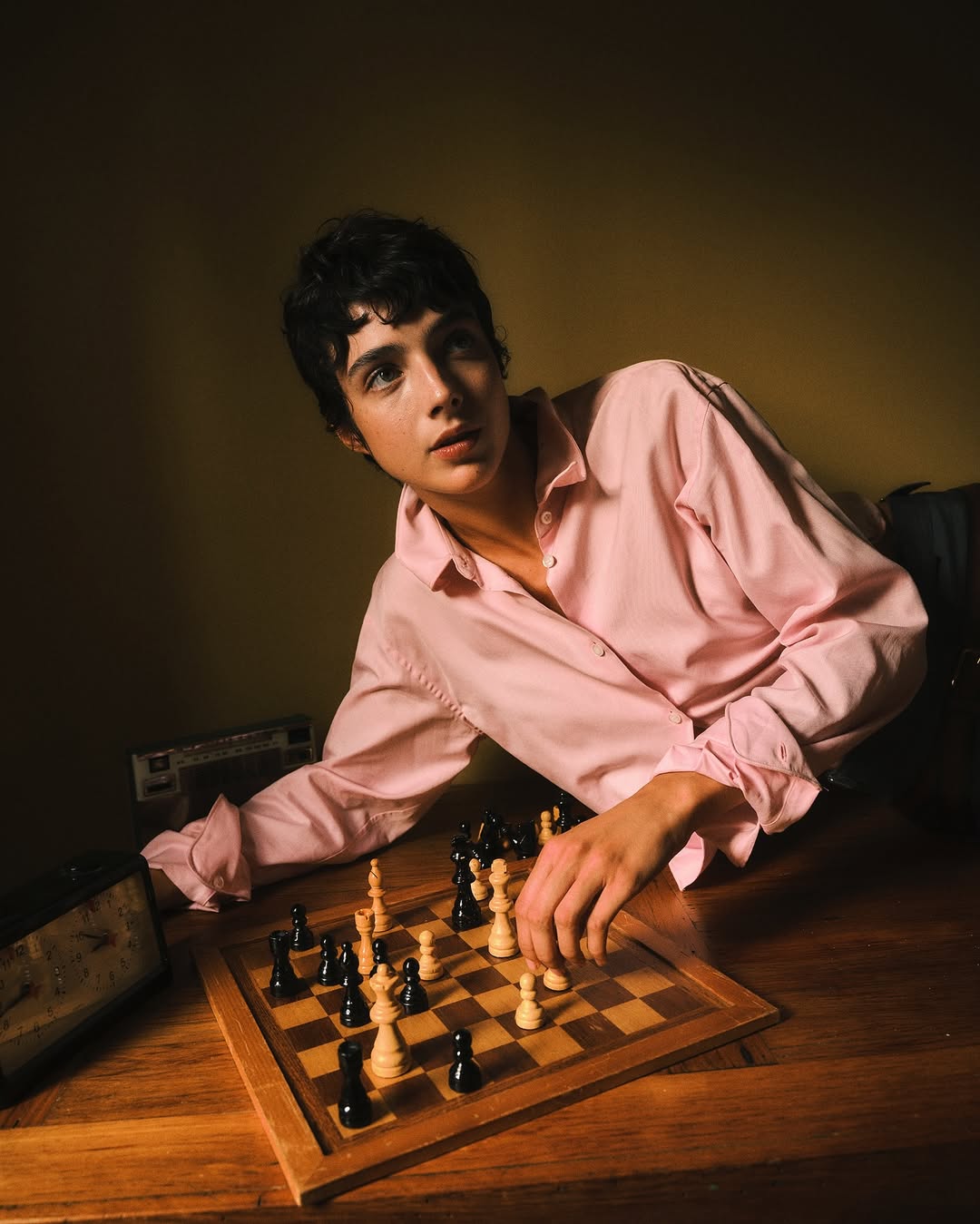 Indoor portrait of a young adult at a chess board with pieces, wearing a pink collared shirt against a subdued background.