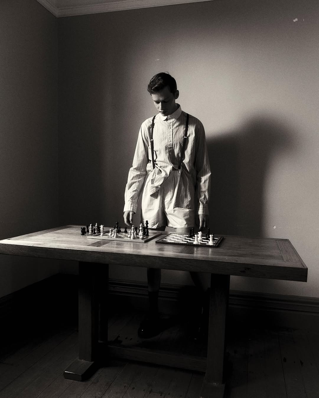 Young man stands indoors at a large wood table with two chessboards. The black and white image has a simple, minimalist aesthetic.