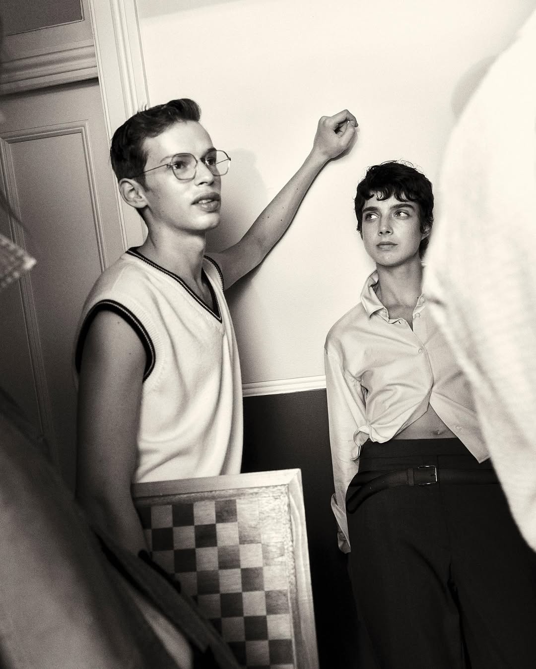Monochrome portrait of a young adult couple posing indoors against a white wall, featuring a checkerboard and vintage aesthetic.