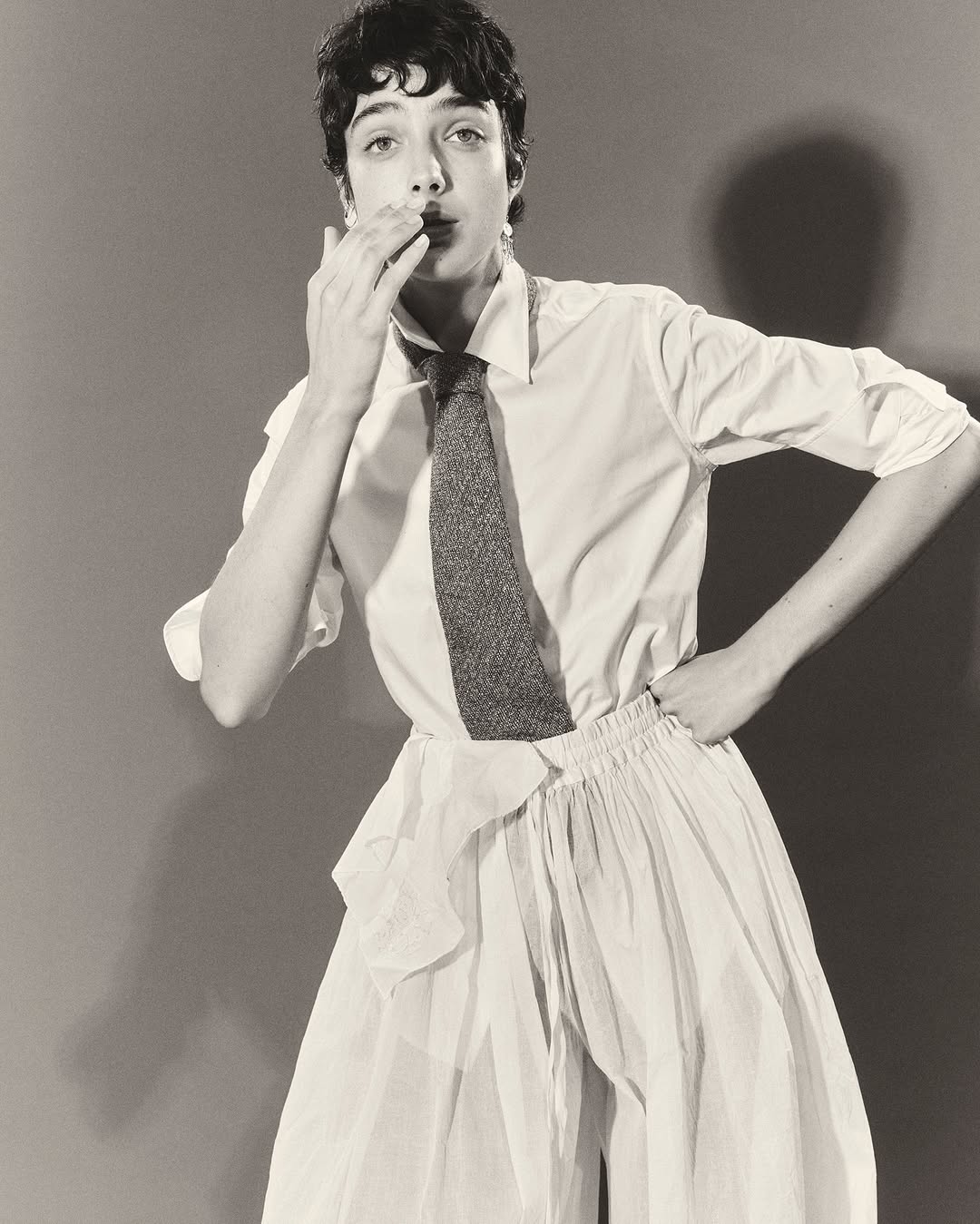 Stylish studio portrait of a young adult in a white shirt and skirt, accessorized with a gray tie, against a neutral background.
