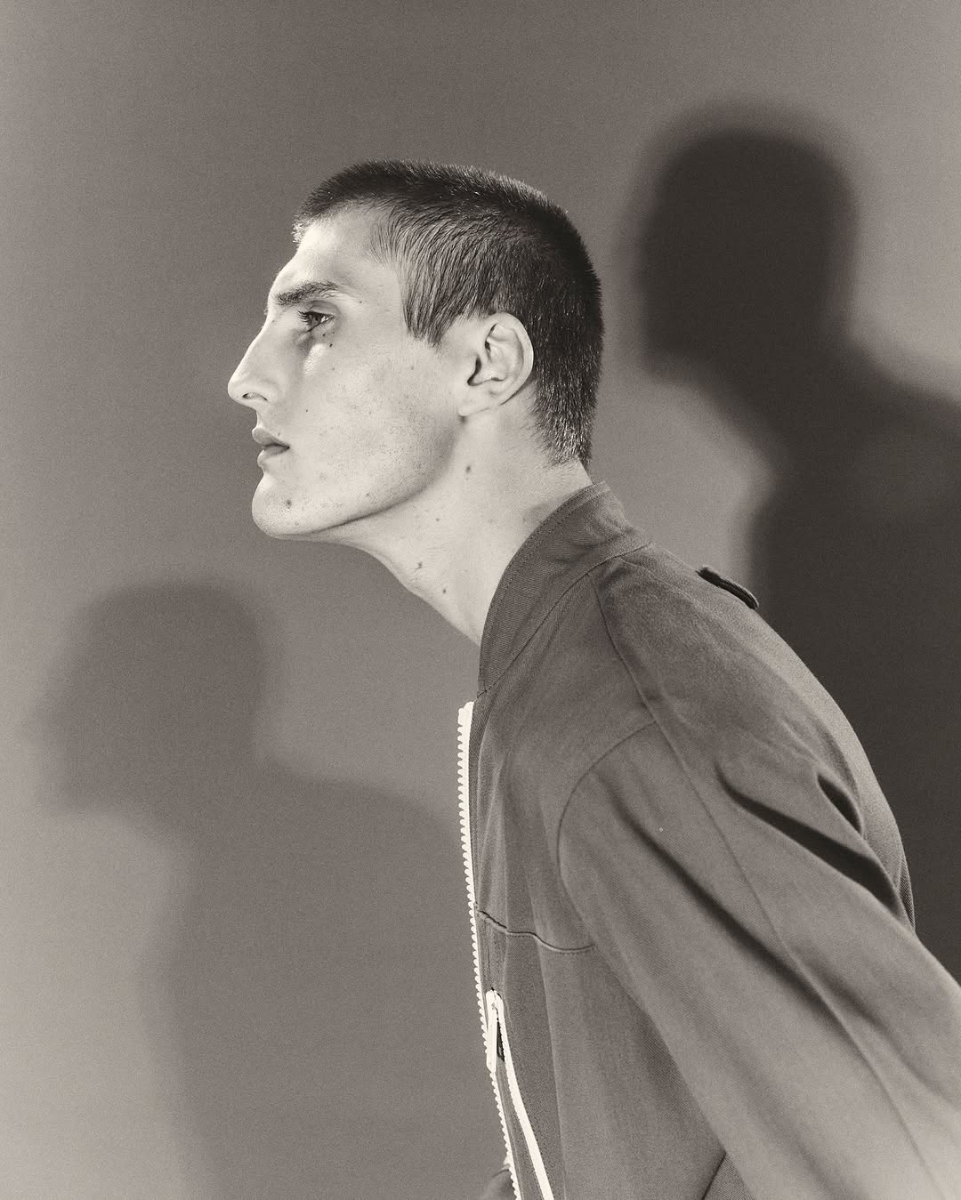 Monochromatic side-view studio portrait of a young adult male with a buzzcut, wearing a bomber jacket against a solid backdrop with a shadow.
