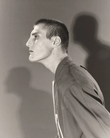 Monochromatic side-view studio portrait of a young adult male with a buzzcut, wearing a bomber jacket against a solid backdrop with a shadow.