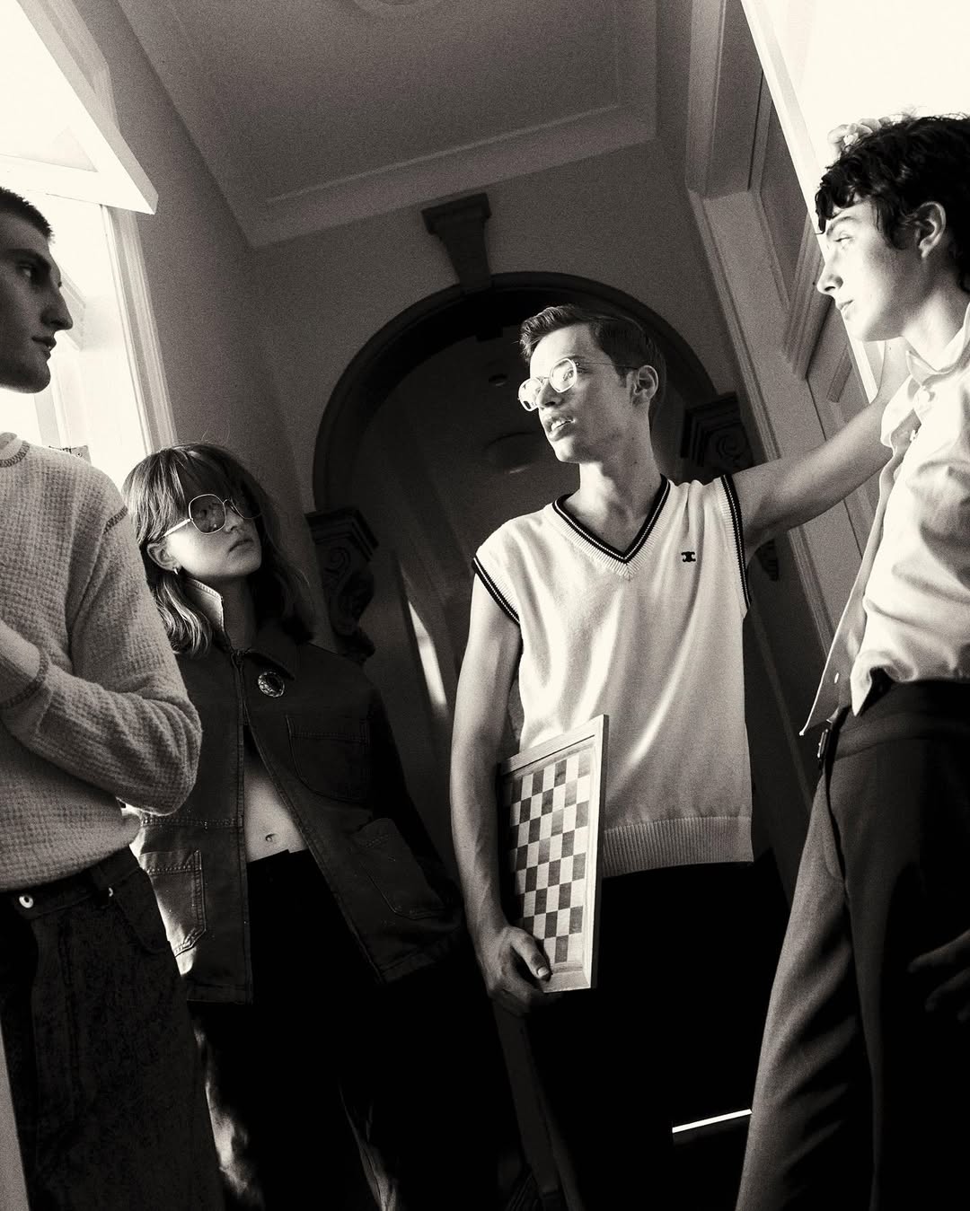 Black and white image of a group of four young adults indoors, one holding a chessboard, standing in a hallway with a doorway in the background.