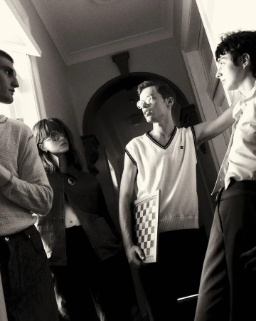 Black and white image of a group of four young adults indoors, one holding a chessboard, standing in a hallway with a doorway in the background.