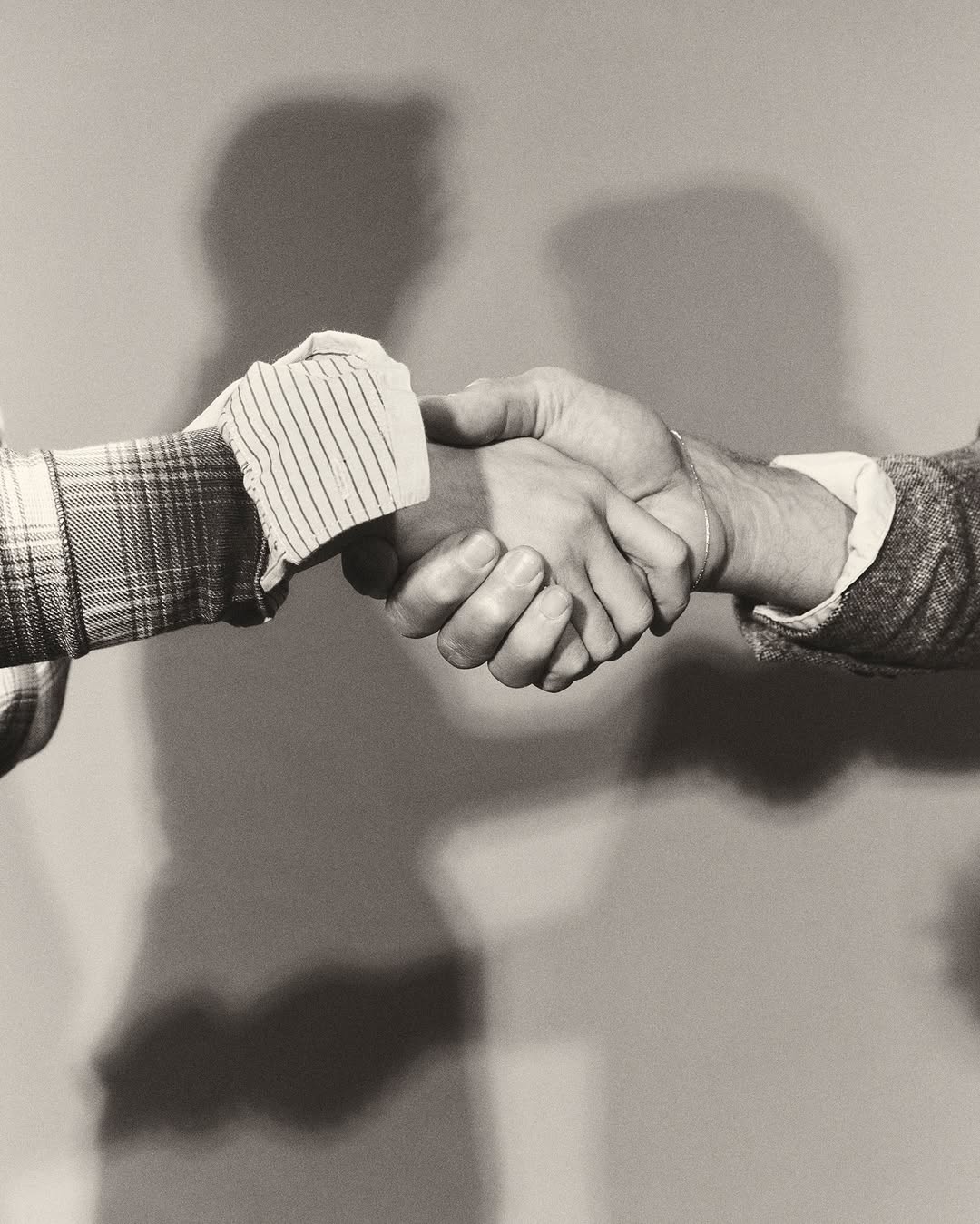 Monochrome image of two people shaking hands, showcasing a close-up of their hands and forearms against a neutral background with shadows.