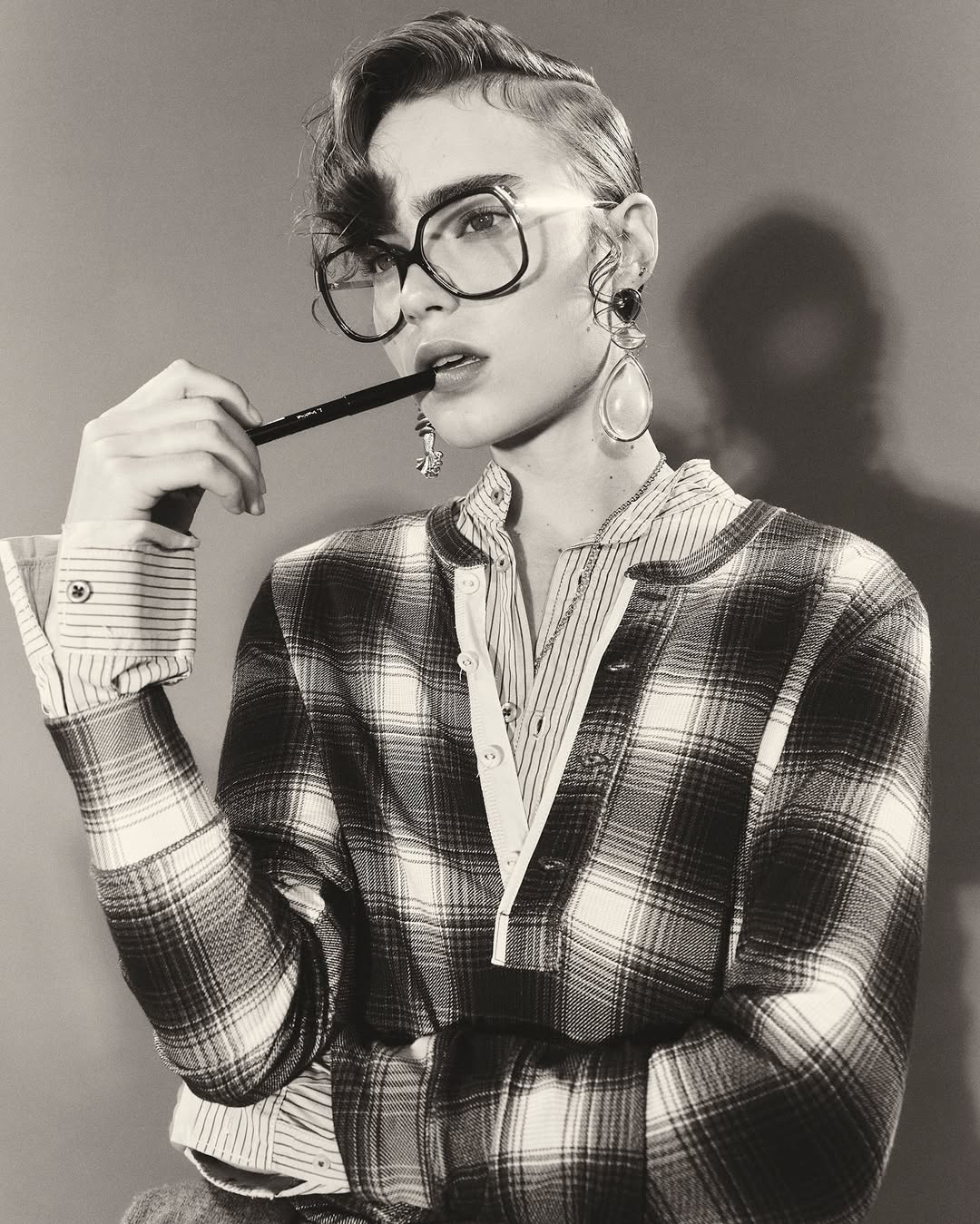 A woman wearing glasses and a plaid shirt poses in a monochrome studio shot, holding a pen to her lips with earring details.