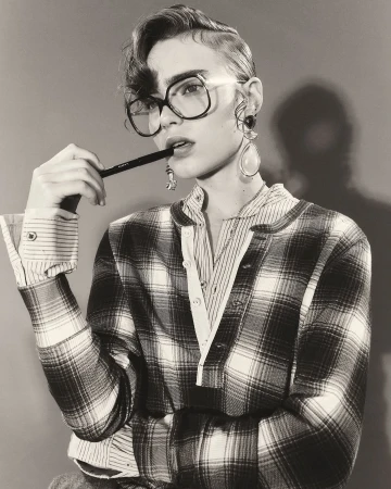A woman wearing glasses and a plaid shirt poses in a monochrome studio shot, holding a pen to her lips with earring details.