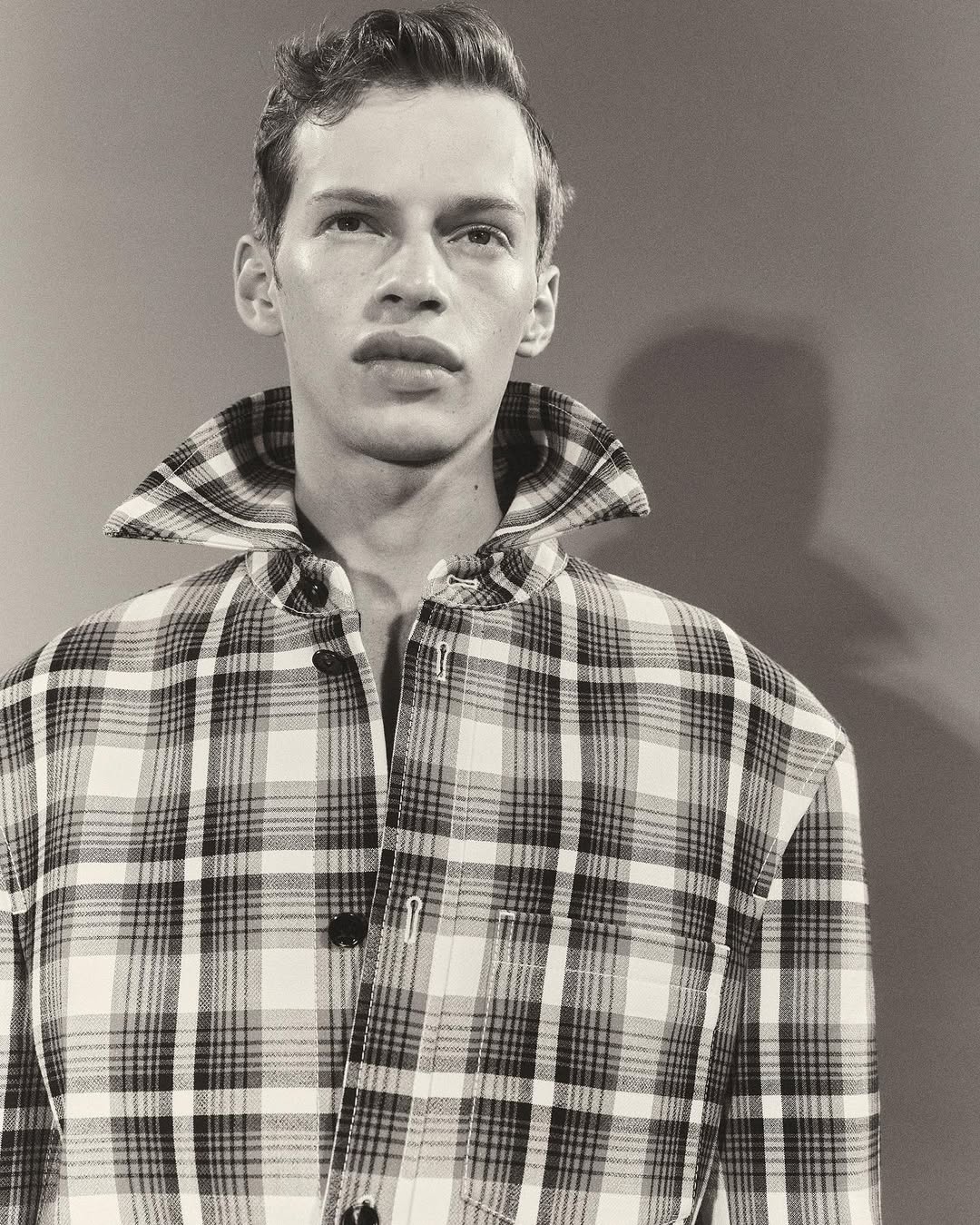 Monochromatic portrait of a young adult male in a plaid jacket with a wide collar, posing against a solid grey background.