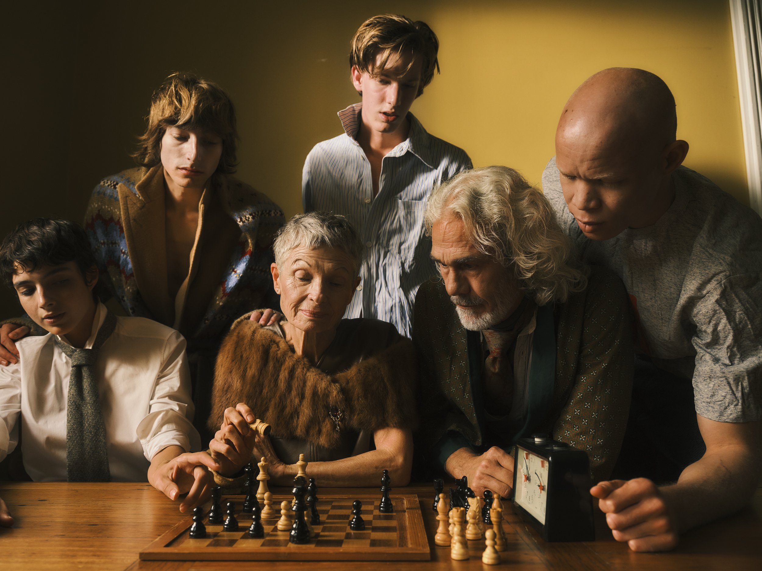 Group of six people, including an elderly woman and man, gathered around a wooden chess board with chess pieces on a yellow background.