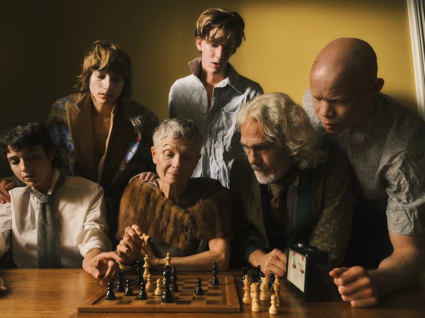 Group of six people, including an elderly woman and man, gathered around a wooden chess board with chess pieces on a yellow background.