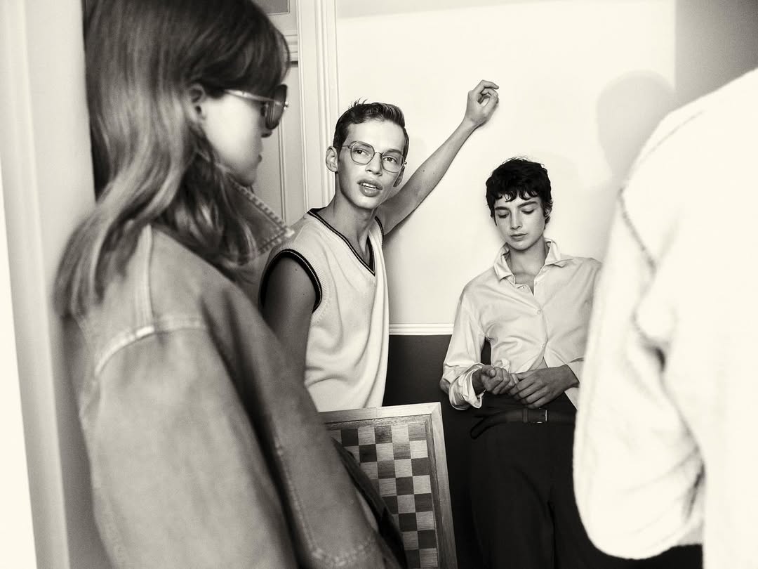 Monochrome indoor shot of three young adults in casual attire, one holding a chessboard, in a simple room with natural lighting.