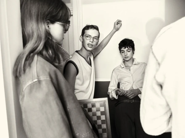 Monochrome indoor shot of three young adults in casual attire, one holding a chessboard, in a simple room with natural lighting.
