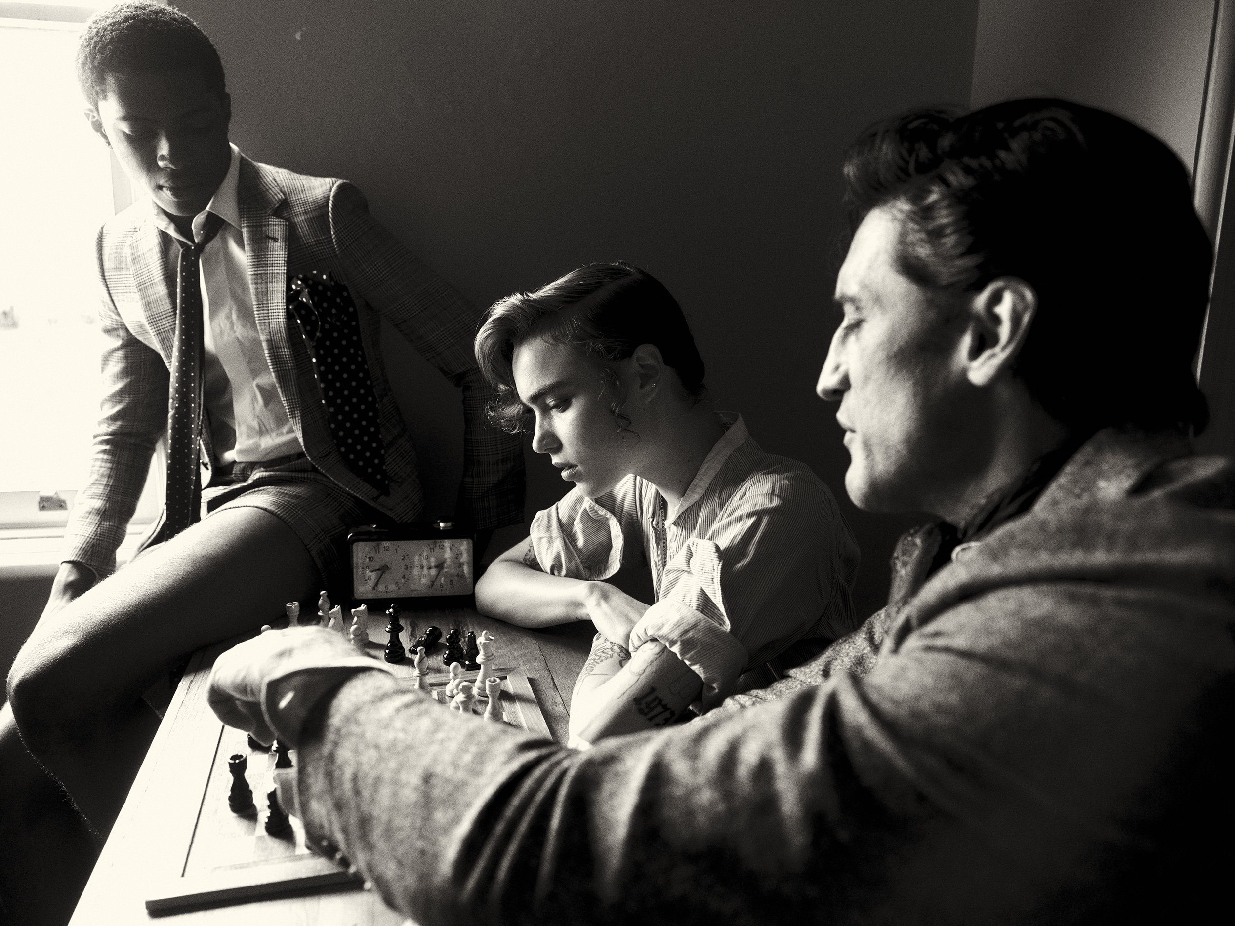 Three men are playing chess at a table indoors in a black and white photograph with a window in the background.
