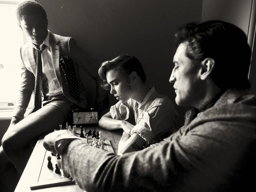 Three men are playing chess at a table indoors in a black and white photograph with a window in the background.