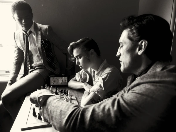 Three men are playing chess at a table indoors in a black and white photograph with a window in the background.