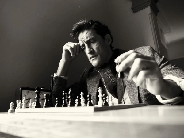 Black-and-white shot of a man contemplating a chess move indoors, with an alarm clock visible in the background.