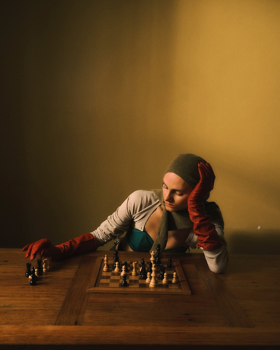 A young woman in a headscarf and gloves sits at a wooden table, contemplating a chessboard with black and white pieces under warm lighting.