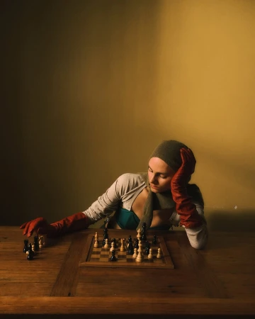 A young woman in a headscarf and gloves sits at a wooden table, contemplating a chessboard with black and white pieces under warm lighting.